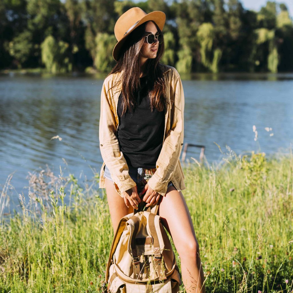 young-stylish-woman-standing-near-river-with-holding-backpack-min